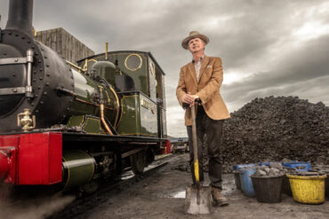 Paul Merton standing next to a steam train and coal pile with shovel in hand
