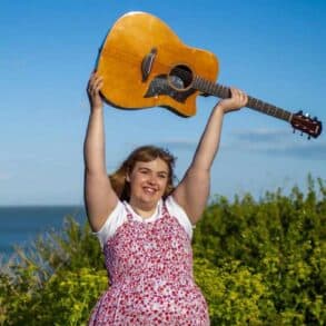 Amy holding her guitar in the air whilst standing in a field by the coast