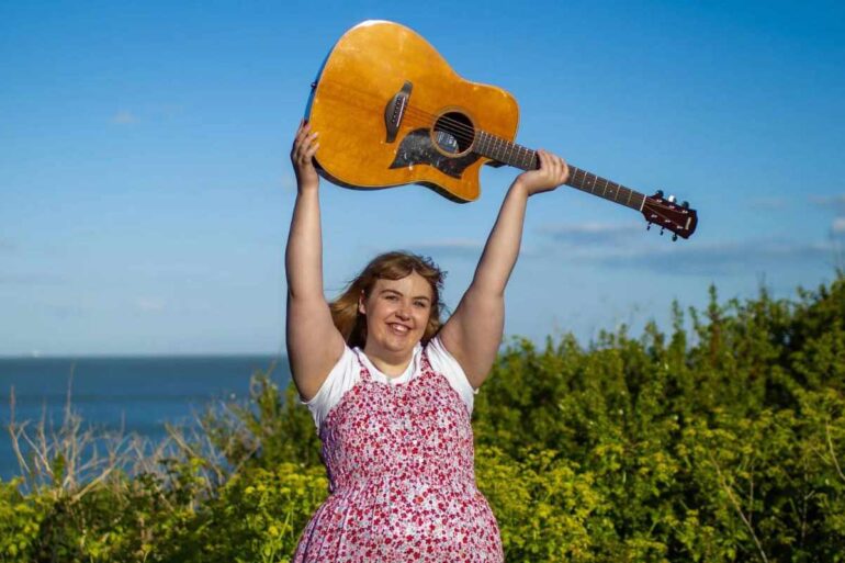 Amy holding her guitar in the air whilst standing in a field by the coast