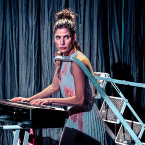 Gabriela Flarys performing Deluge on stage - playing an organ whilst having her head through a stepladder