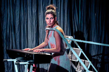 Gabriela Flarys performing Deluge on stage - playing an organ whilst having her head through a stepladder