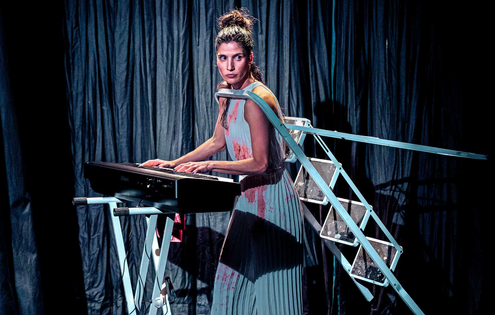 Gabriela Flarys performing Deluge on stage - playing an organ whilst having her head through a stepladder