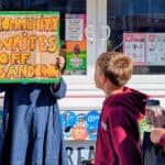 Person holding up a sign that reads' Community writes off Sandown' with school children walking past