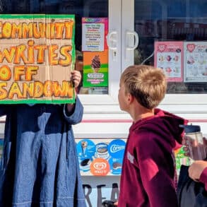 Person holding up a sign that reads' Community writes off Sandown' with school children walking past
