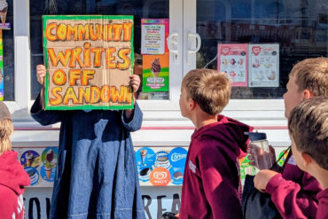 Person holding up a sign that reads' Community writes off Sandown' with school children walking past