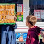 Person holding up a sign that reads' Community writes off Sandown' with school children walking past
