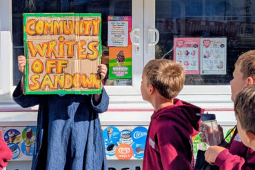 Person holding up a sign that reads' Community writes off Sandown' with school children walking past