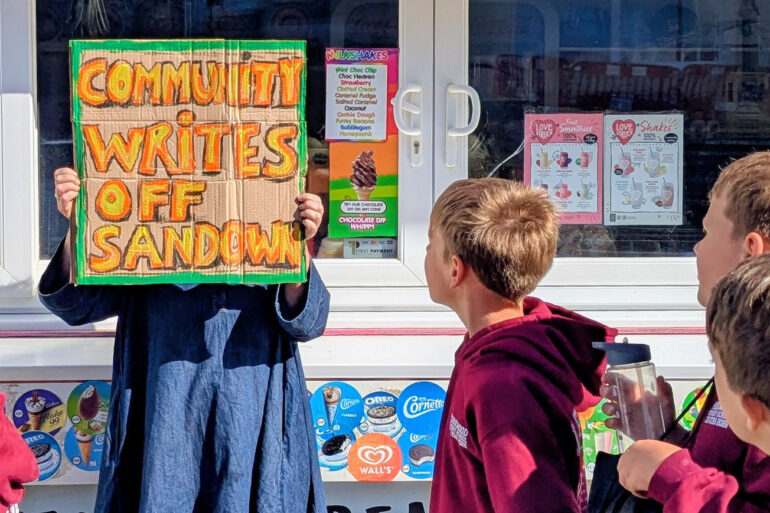 Person holding up a sign that reads' Community writes off Sandown' with school children walking past