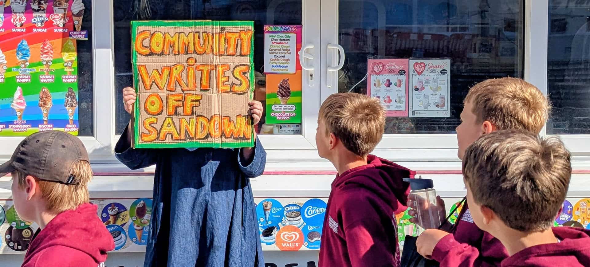 Person holding up a sign that reads' Community writes off Sandown' with school children walking past