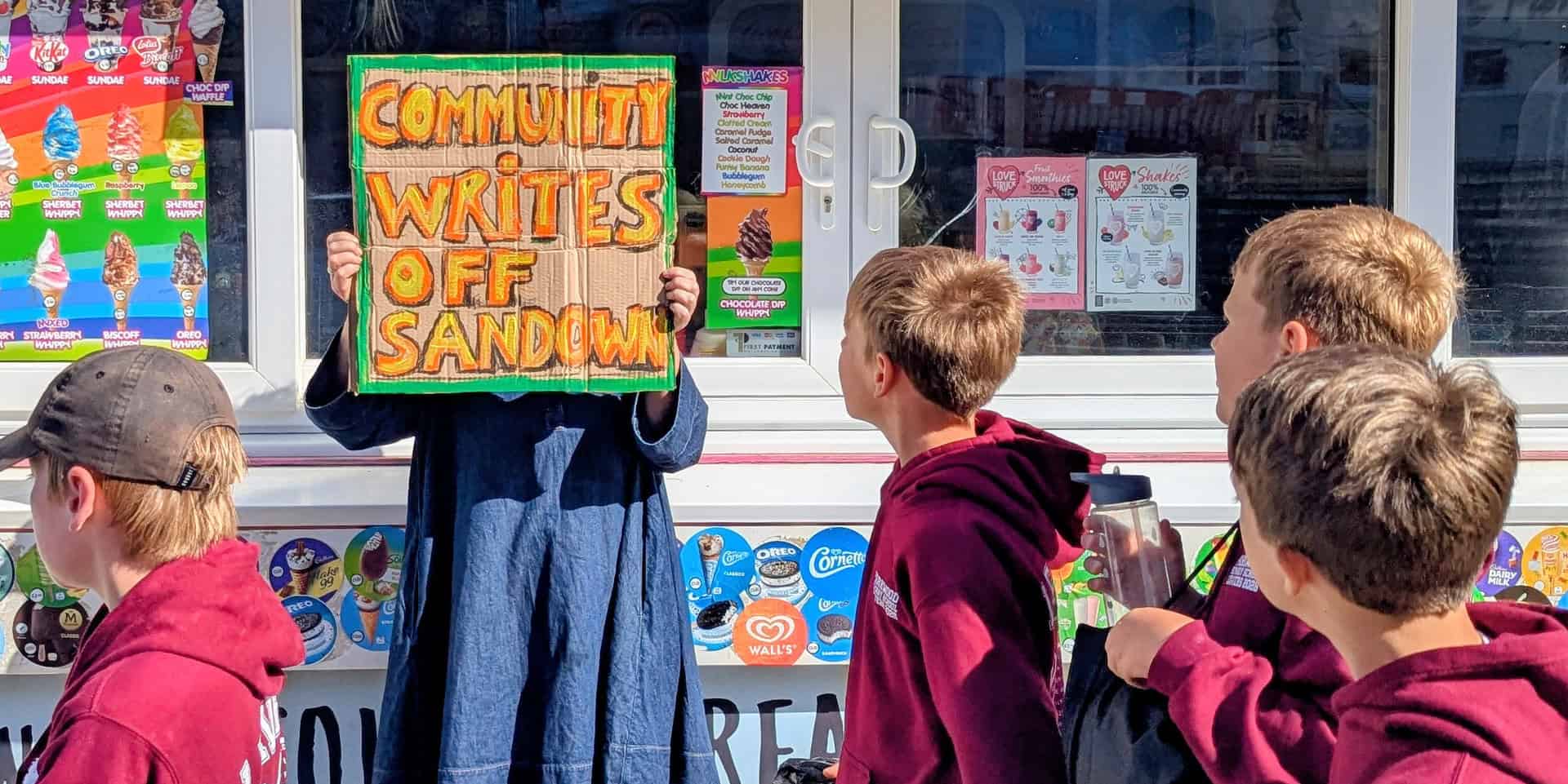 Person holding up a sign that reads' Community writes off Sandown' with school children walking past