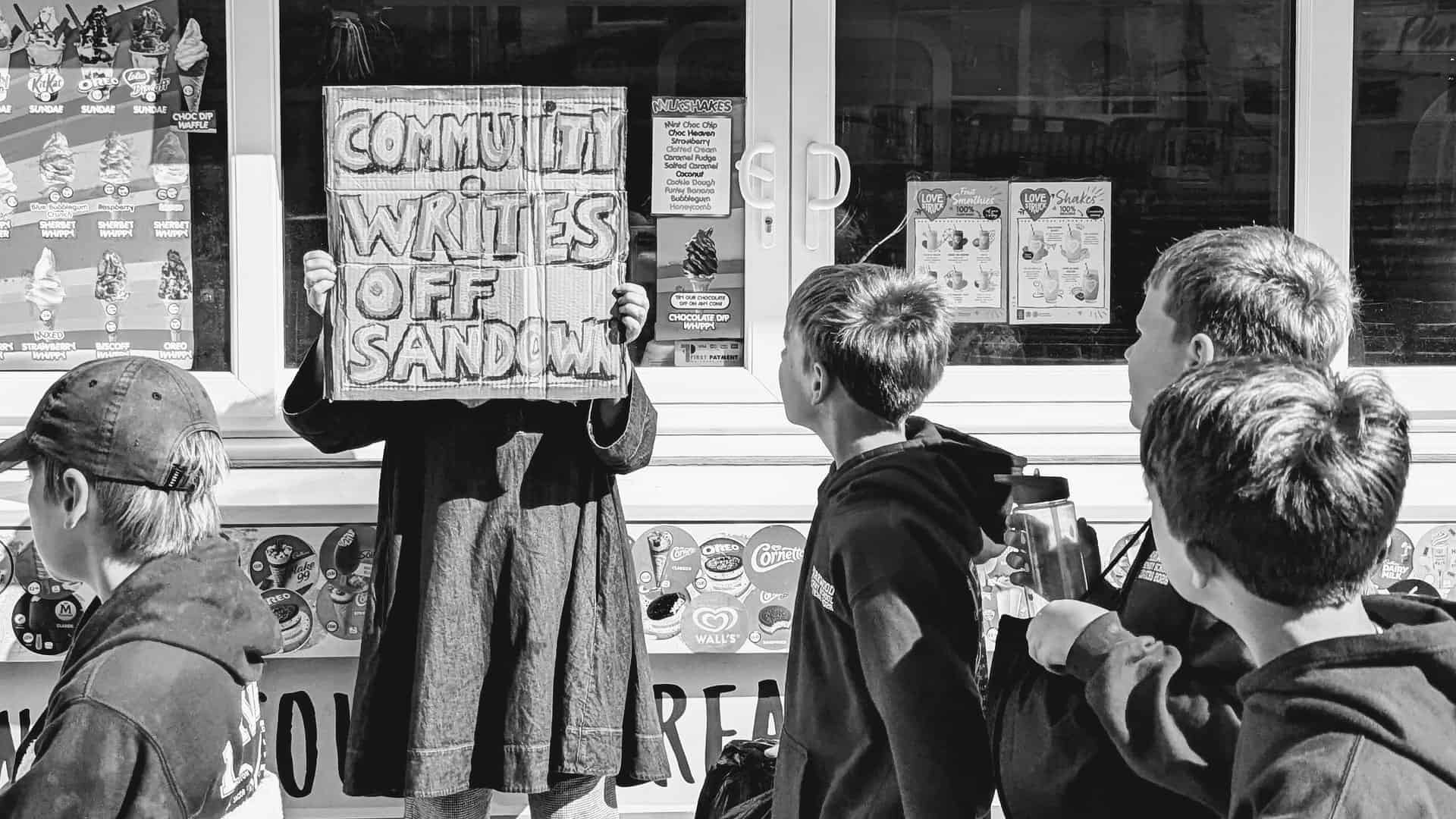Person holding up a sign that reads' Community writes off Sandown' with school children walking past