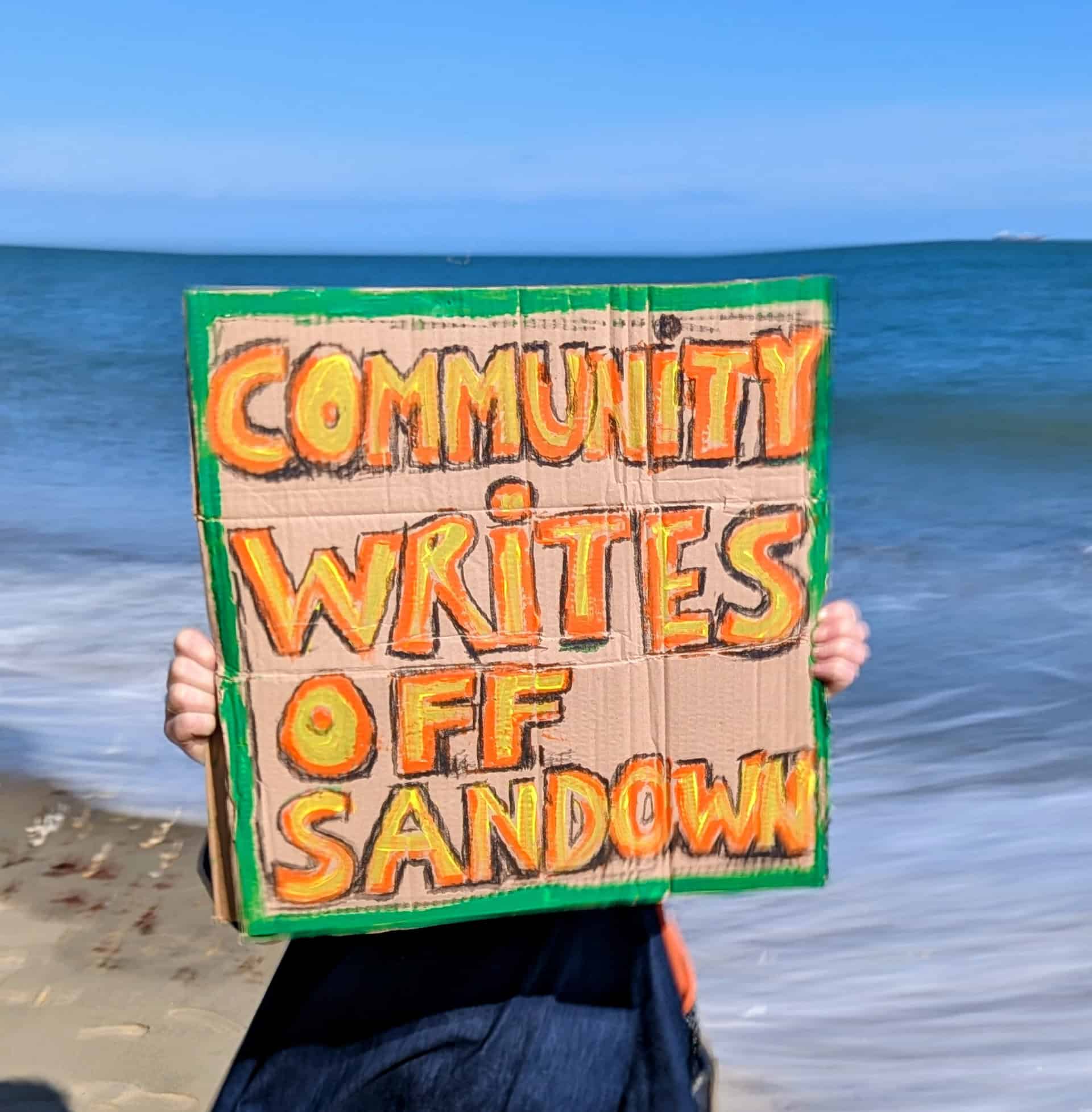 Person on the beach in front of a wonky horizon holding up a sign that reads' Community writes off Sandown'