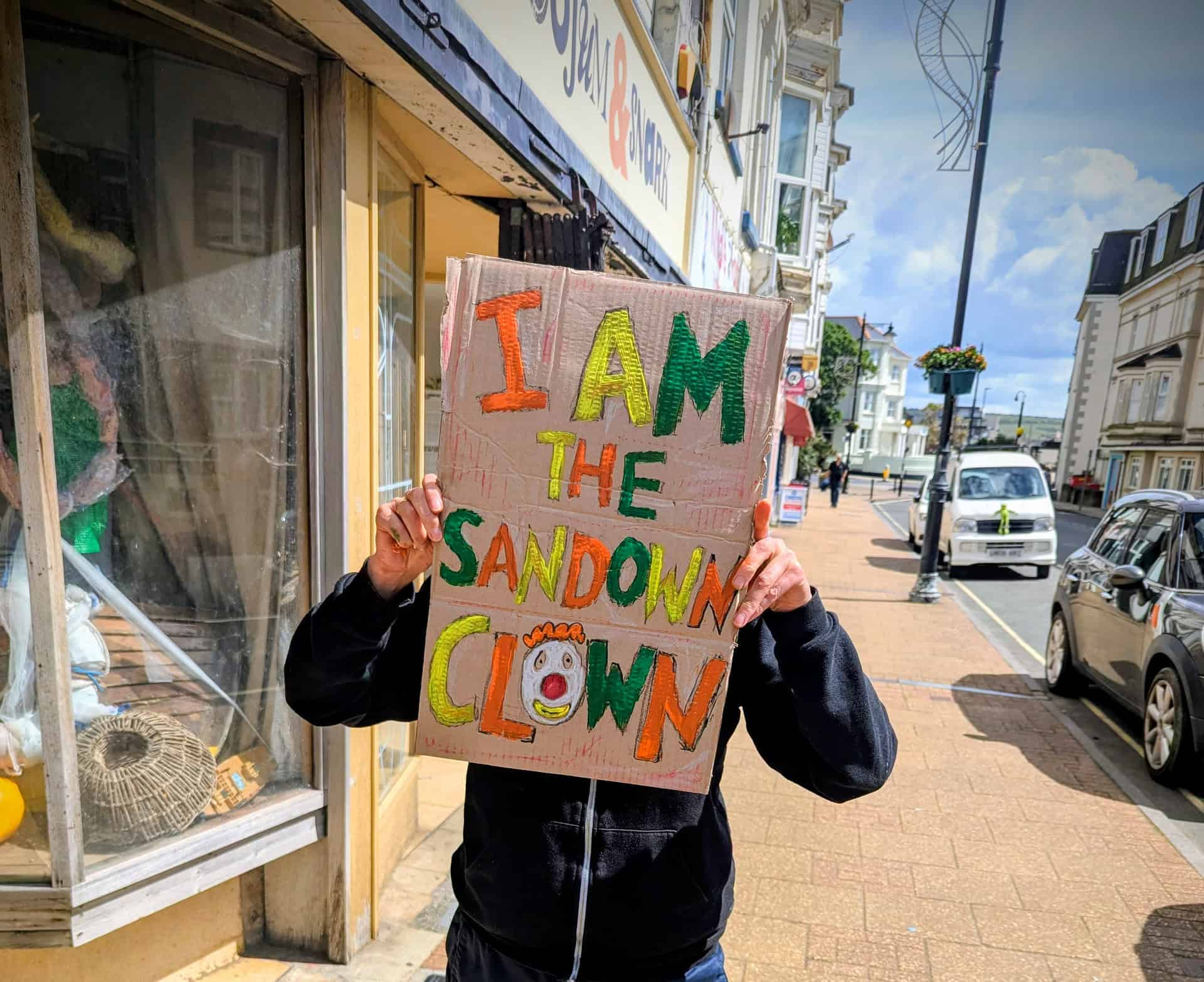 Person holding a sign that reads "i am the sandown clown"