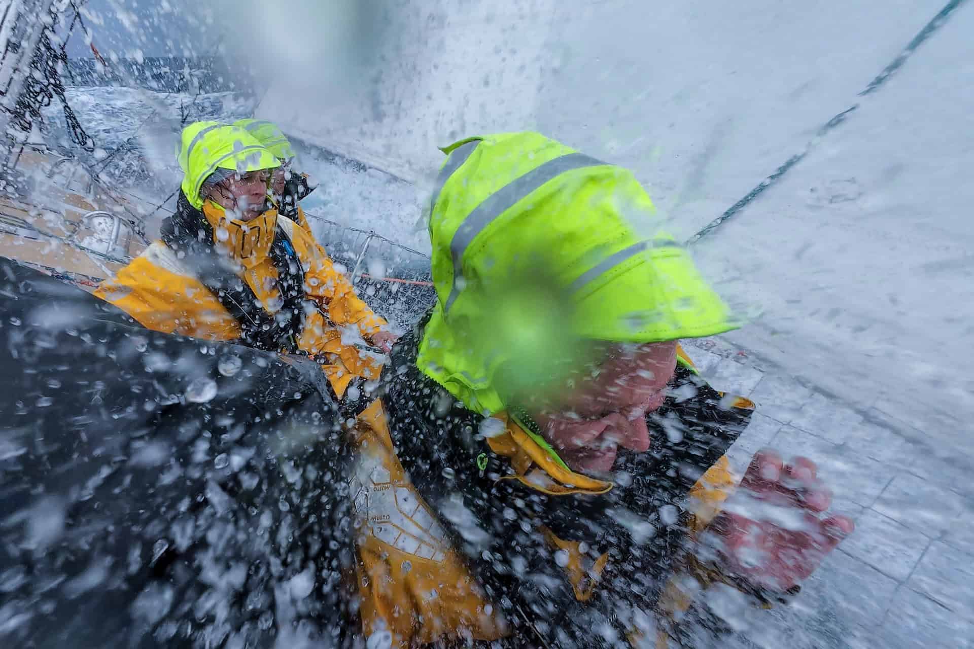 Two sailors on board the yacht during stormy conditions, being sprayed with sea water