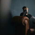 a man holding his head in his hands whilst sat on a leather chesterfield sofa