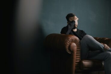 a man holding his head in his hands whilst sat on a leather chesterfield sofa