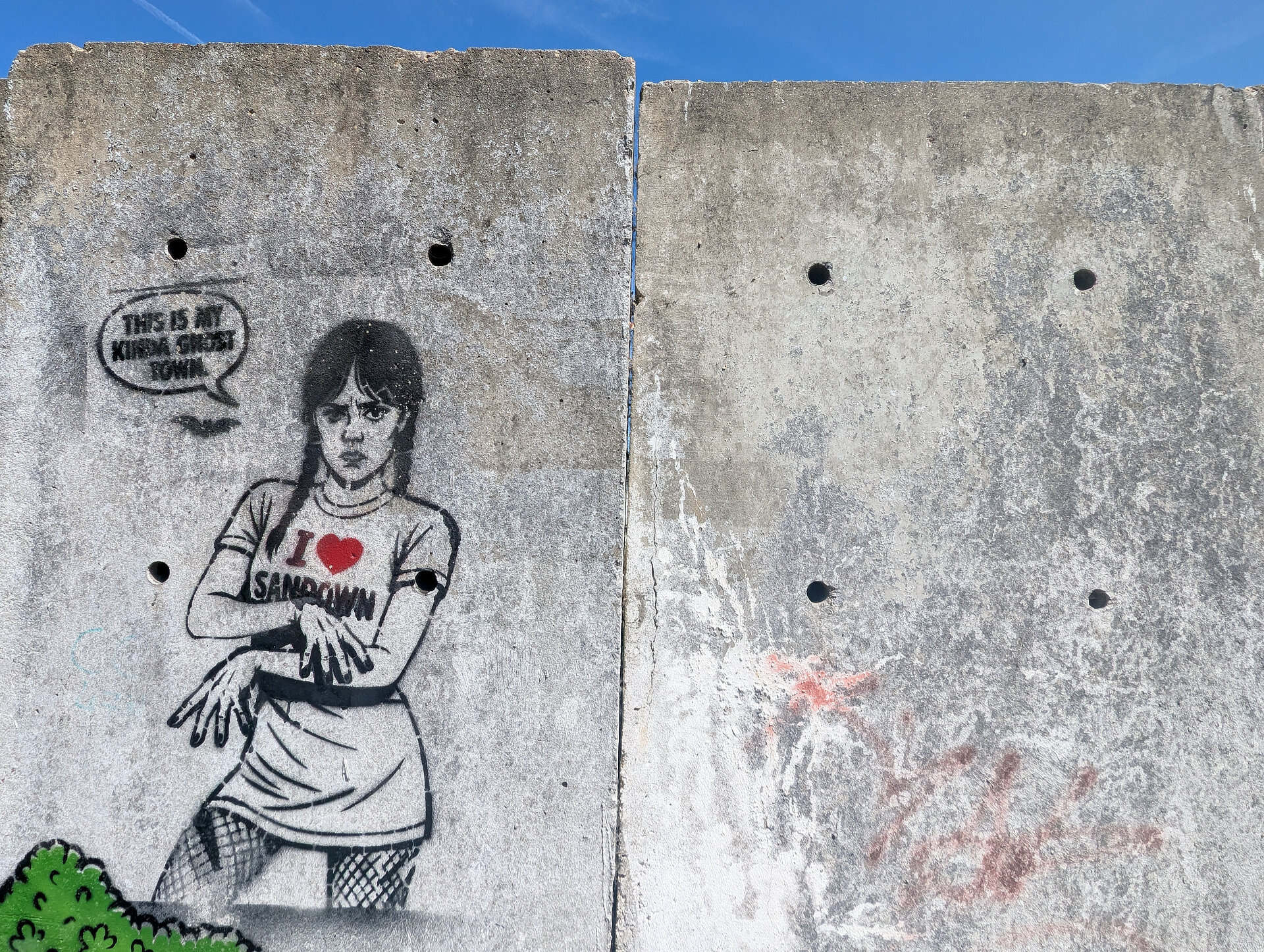 Stencil art on a concrete wall of the character Wednesday wearing a I Love Sandown T-shirt and speech bubble that says, 'this is my kinda ghost town'