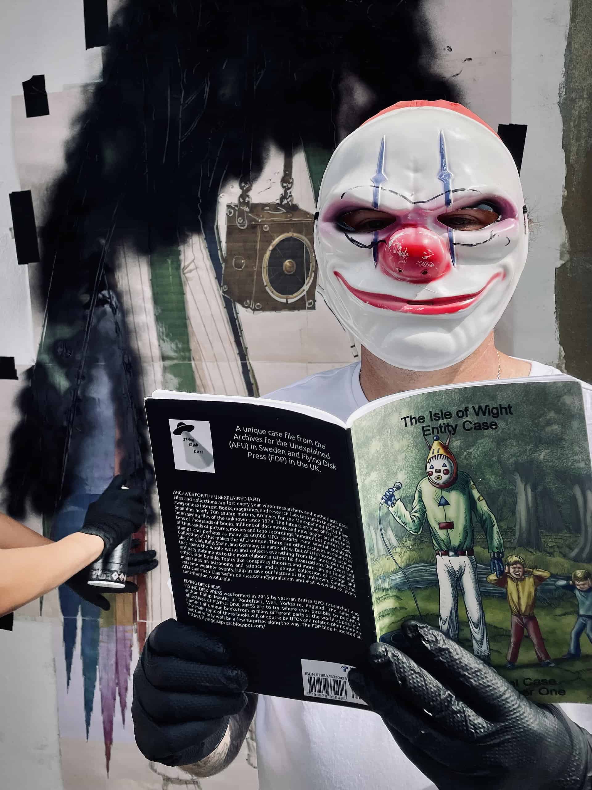 Masked graffiti artists holding Sandown Clown book, whilst artist sprays paint in the background