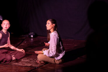 Two dancers sat crossed legged opposite each other on a stage
