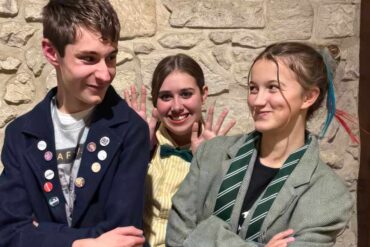 the young Lovestruck Alley performers standing side to side with their arms crossed and one standing behind the other two with waving hands