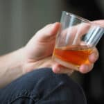Man in jeans sitting down on the ground with a glass of whisky in his hands