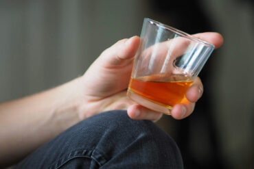 Man in jeans sitting down on the ground with a glass of whisky in his hands