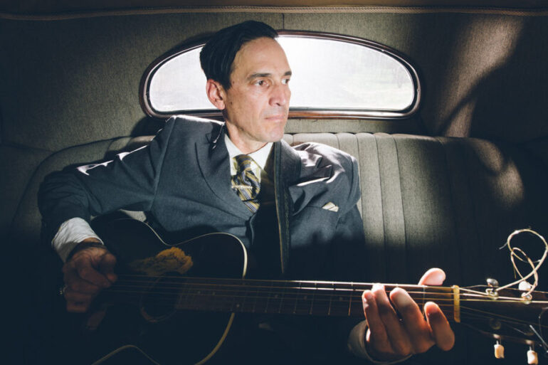 Man in uniform sitting in the backseat of a vintage car with his guitar